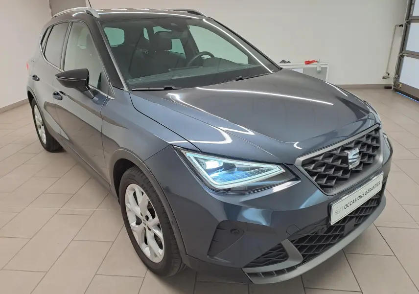 SEAT Arona 2024 gris magnétique avec toit noir, vue 3/4 avant droit, phares allumés en intérieur showroom.