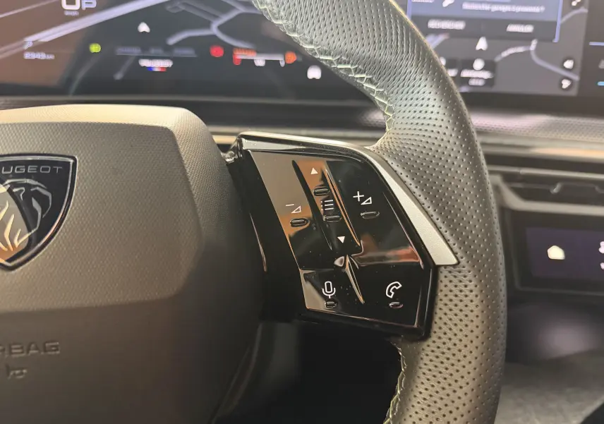 Gros plan sur le volant en cuir perforé de la Peugeot 3008 Hybrid 145 GT, avec commandes audio et affichage numérique en arrière-plan.