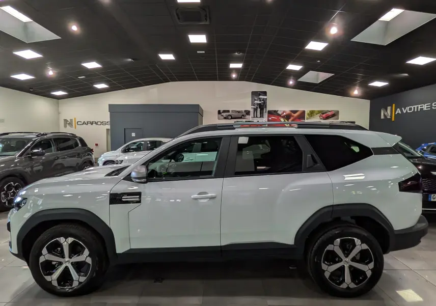 Vue latérale gauche du Dacia Bigster blanc glacier 2026 en version Mild Hybrid 140 CH Journey dans un showroom.
