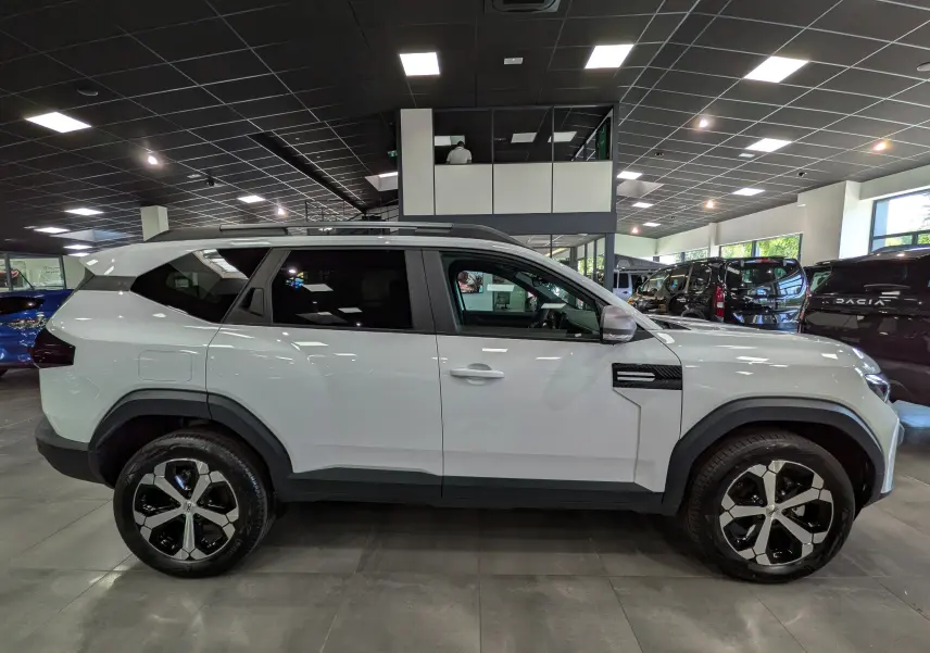 Vue de profil côté gauche du SUV blanc glacier Dacia Bigster Mild Hybrid 140 CH Journey avec jantes bicolores en concession.