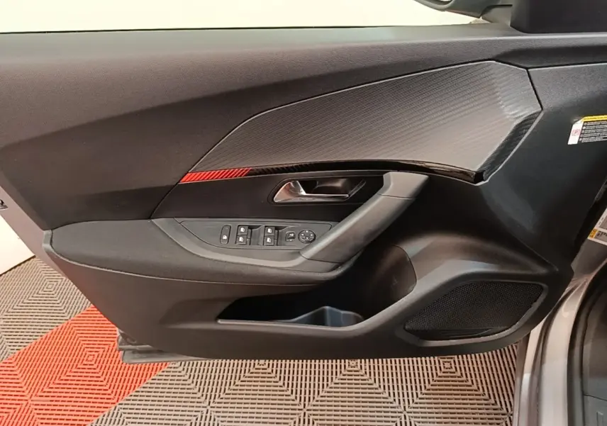 Vue intérieure côté gauche de la porte avant gris clair du Peugeot E-2008 électrique avec insert carbone et bande rouge décorative.
