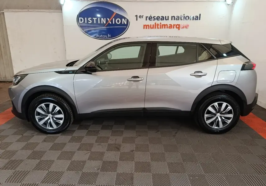 Vue latérale droite d’un Peugeot E-2008 électrique Active Pack gris clair dans un showroom intérieur.