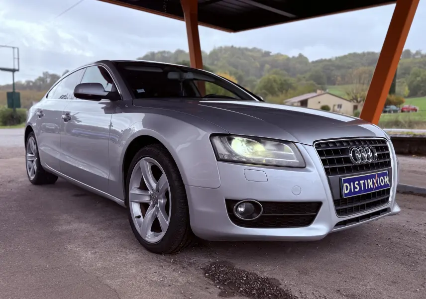 Audi A5 gris en 3/4 avant droit sous abri, avec phares allumés et jantes alliage cinq branches.