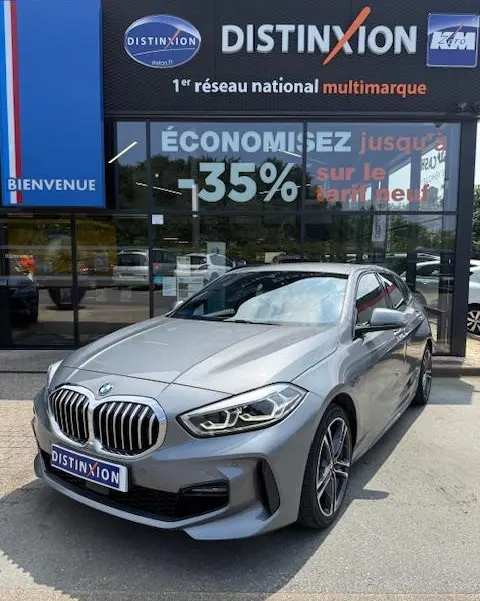 BMW Série 1 118d Edition Sport gris foncé vue 3/4 avant, avec calandre et phares LED distinctifs, devant un showroom multimarque.