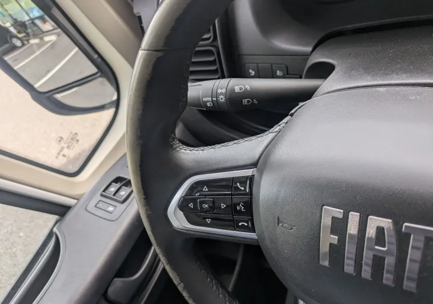 Gros plan sur le volant noir du Fiat Ducato Fourgon 2025 avec commandes intégrées côté gauche et rétroviseur extérieur visible.