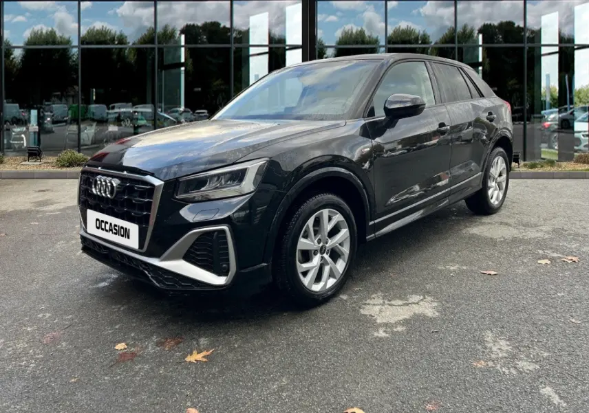 Audi Q2 noir vue 3/4 avant droit, avec calandre distinctive et jantes argentées sur parking devant vitres réfléchissantes.