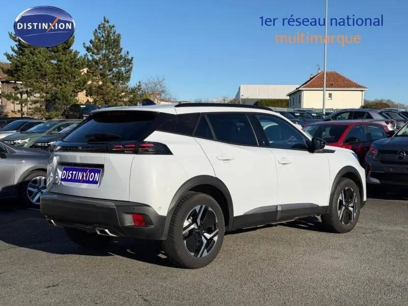 Vue 3/4 arrière droite d'un Peugeot 2008 blanc avec barres de toit et jantes aluminium sur parking.