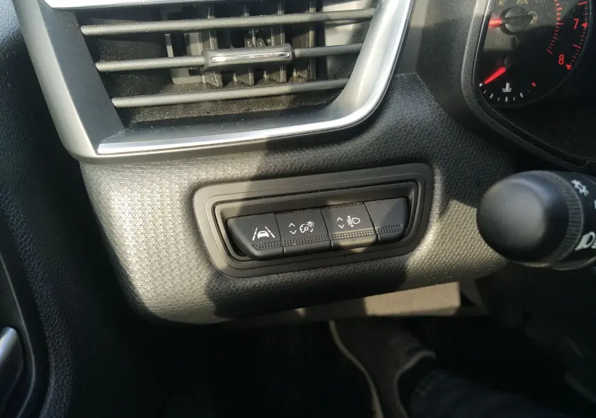 Détail du tableau de bord gris de la Renault Clio V 2023 montrant les boutons de réglage des aides à la conduite et des phares.