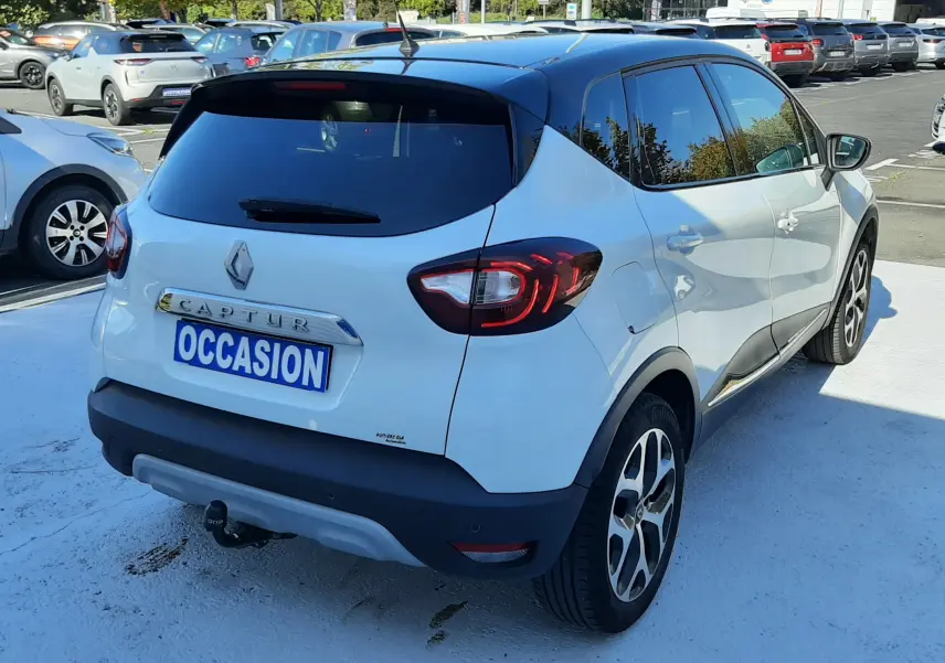 Vue 3/4 arrière droite d'un Renault Captur blanc nacré avec toit noir, équipé d'une attache remorque sur parking.