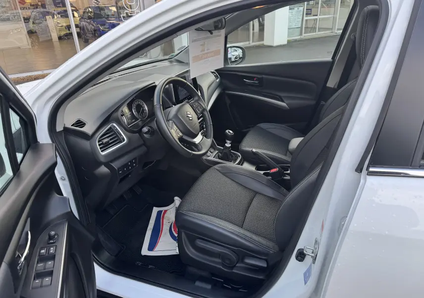 Intérieur noir du Suzuki S-Cross blanc 2023, vue côté conducteur avec volant, sièges et tableau de bord visibles.
