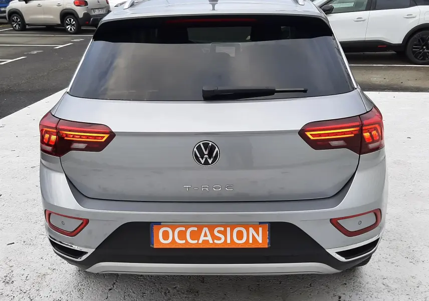 Vue arrière d'un Volkswagen T-Roc gris pyrite avec feux allumés et plaque orange "OCCASION" sur parking.