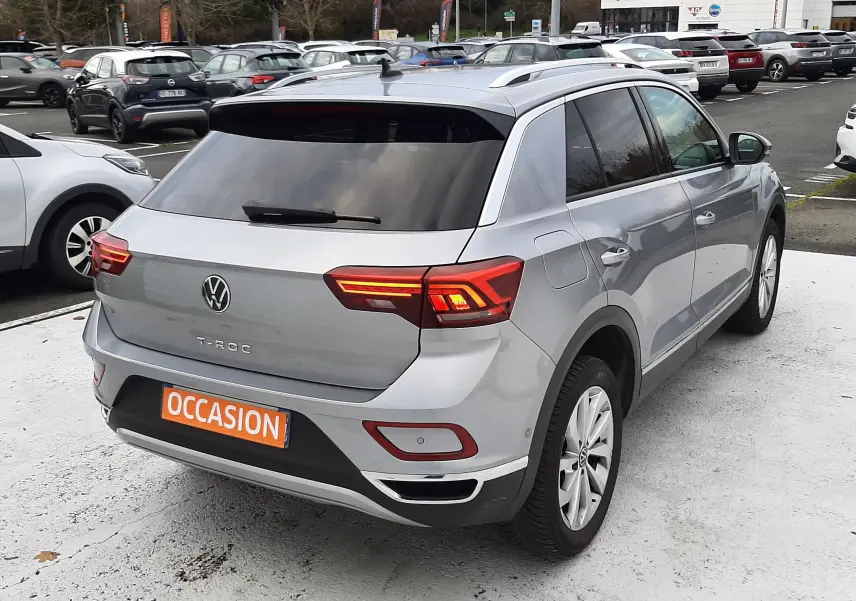 Volkswagen T-Roc gris pyrite vu de l'arrière droit, feux LED allumés, vitres arrière surteintées, sur parking extérieur.