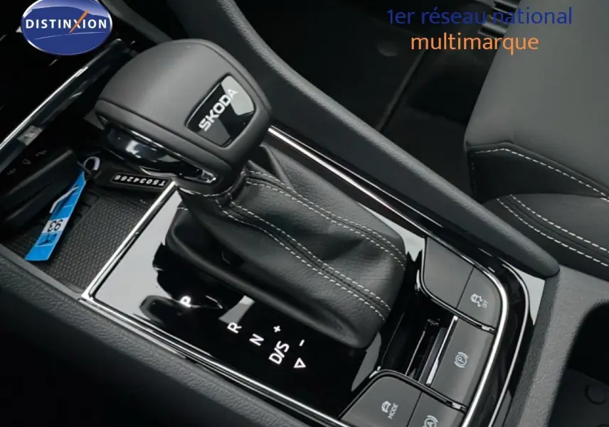 Le levier de vitesse automatique noir avec logo SKODA, entouré de commandes et coutures blanches sur console noire brillante.