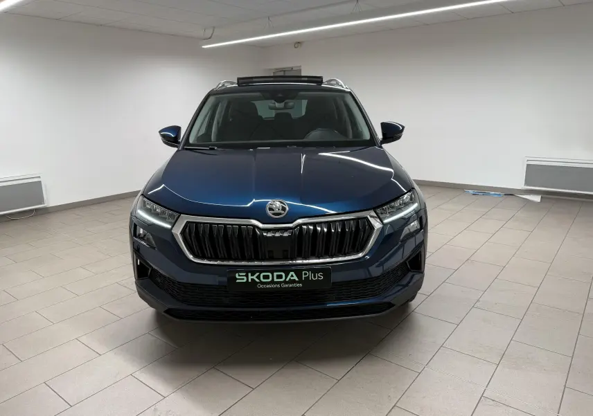Vue frontale d'un Skoda Karoq 2023 bleu lave avec calandre chromée et toit ouvrant en intérieur showroom.