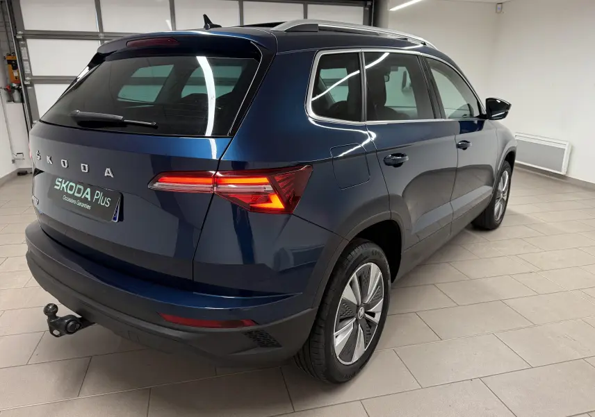 Vue 3/4 arrière droite d'un Skoda Karoq bleu lave avec attelage et feux arrière allumés en intérieur.