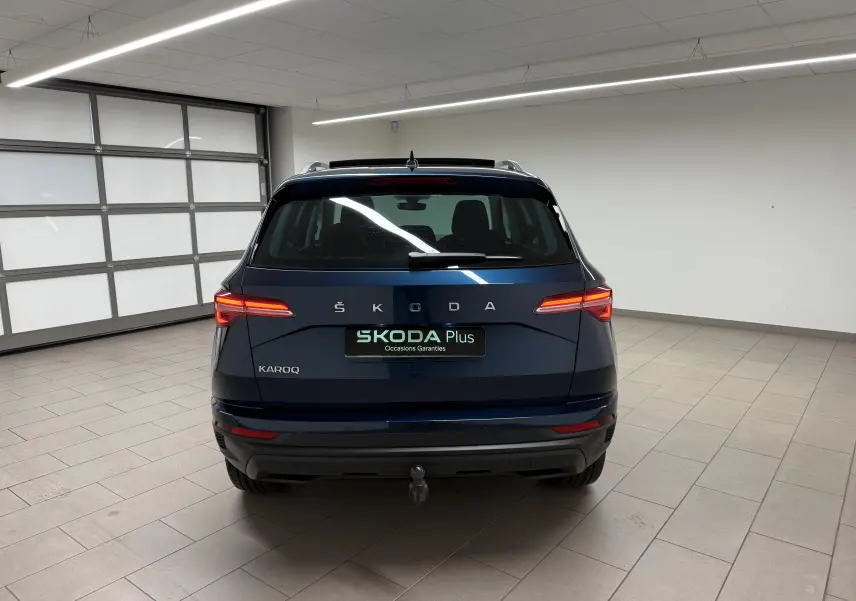 Vue arrière d'un Skoda Karoq bleu lave 2023 avec toit ouvrant et attelage dans un garage lumineux.
