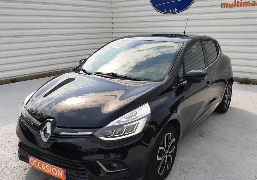 Renault Clio IV noire vue 3/4 avant droit, avec phares LED et jantes alliage bicolores, stationnée devant un mur blanc.