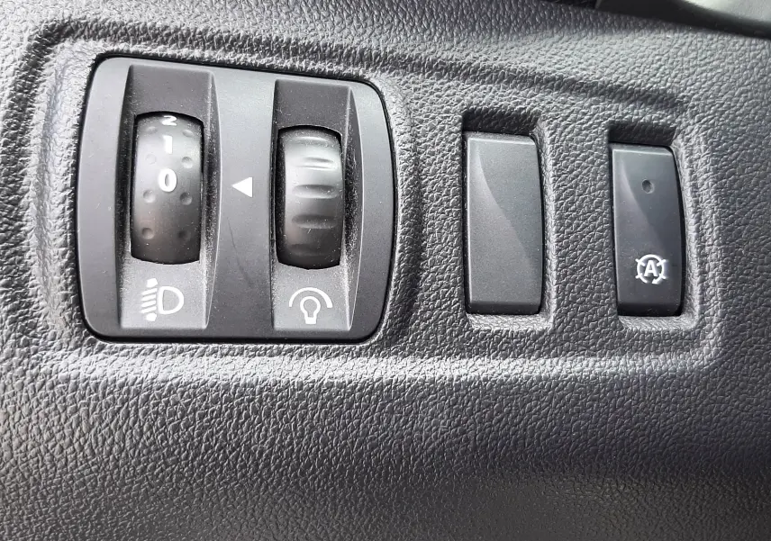 Gros plan sur les commandes noires de réglage des phares et fonctions auxiliaires du tableau de bord d'une Renault Clio IV noire.
