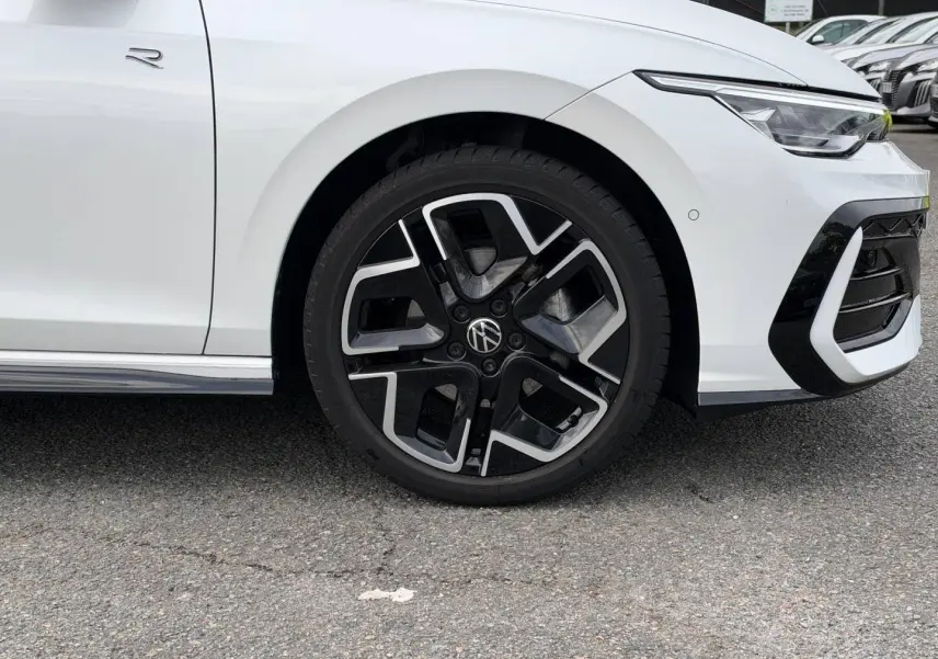 Gros plan sur la roue avant droite et partie avant blanche nacrée d'une Volkswagen Golf R-Line 2025 avec jante alu noire et argent.