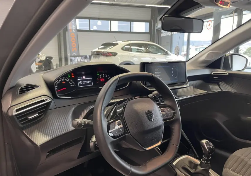 Vue intérieure du poste de conduite de la Peugeot 208 gris clair 2024, volant cuir et écran tactile central visible.