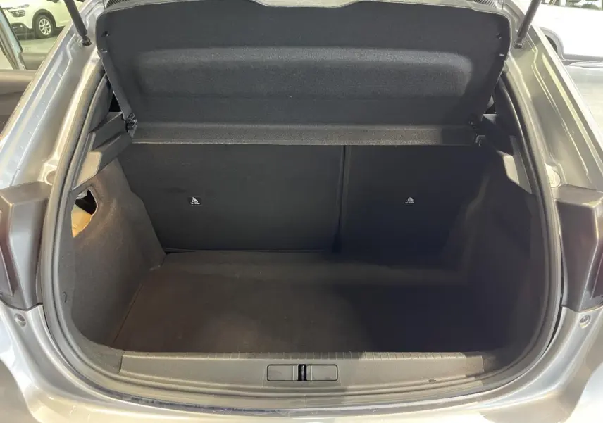 Coffre ouvert vu de l'arrière d'une Peugeot 208 gris clair, montrant l'espace de rangement vide et la trappe de compartiment.