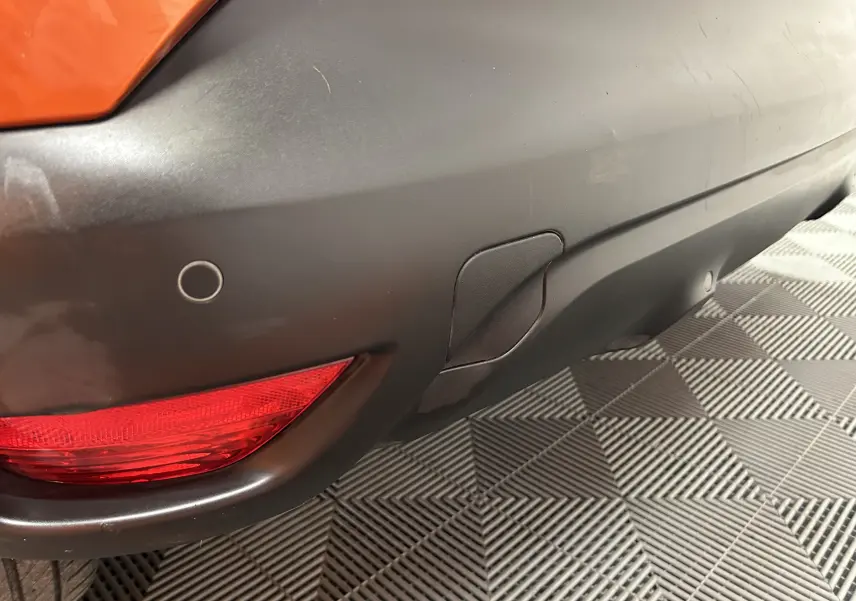Gros plan sur le pare-chocs arrière droit noir mat du Renault Captur orange, avec feu rouge et capteur de radar de recul visible.