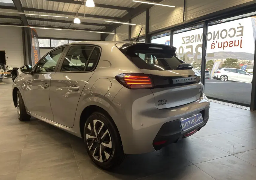 Vue 3/4 arrière droite d'une Peugeot 208 2024 gris clair avec feux arrière à LED et jantes stylisées en concession.
