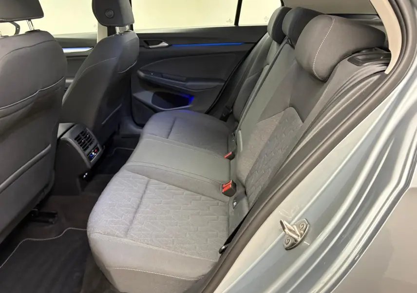 Vue intérieure arrière droite de la Volkswagen Golf 2.0 TDI 2025 en reflet d'argent, sièges tissu gris et éclairage d'ambiance bleu.