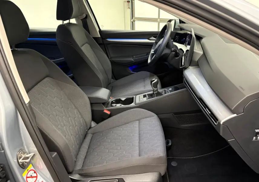 Vue intérieure côté conducteur de la Volkswagen Golf 2.0 TDI 2025 en reflet d'argent, avec sellerie tissu et éclairage d'ambiance bleu.