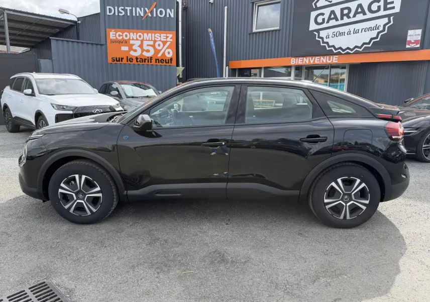 Profil droit d'une Citroën C4 électrique noire 2025 avec inserts noir brillant, stationnée devant un garage.