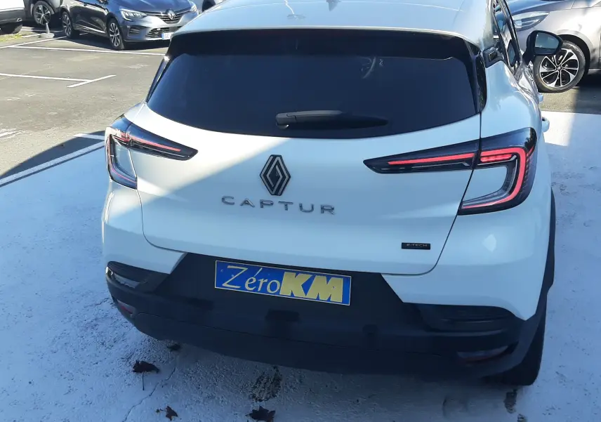 Vue arrière d'un Renault Captur blanc nacré avec toit gris Highland, feux LED allumés et plaque Zéro KM.