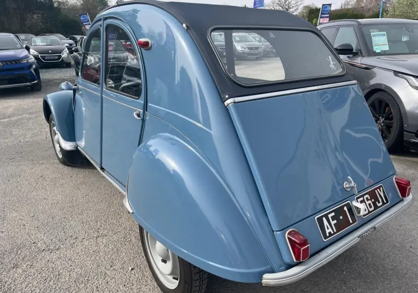 Vue 3/4 arrière gauche d'une Citroën 2CV bleu clair de 1960 avec toit souple noir et plaques d'immatriculation noires.