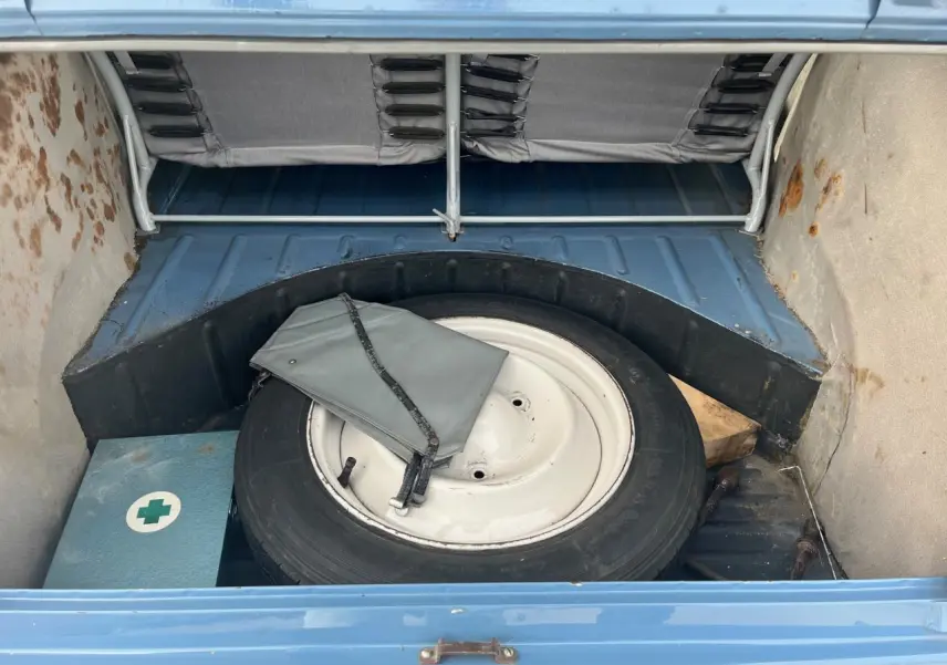 Coffre ouvert d'une Citroën 2 CV bleu clair de 1960, montrant la roue de secours et une trousse de secours.