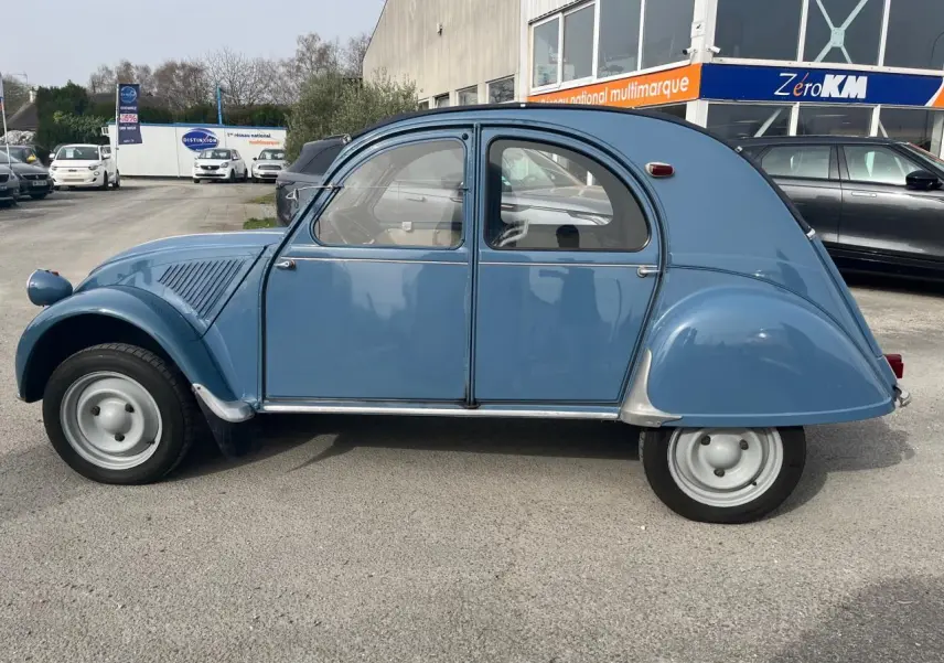 Profil droit d'une Citroën 2 CV bleu clair de 1960, avec ses roues étroites et sa carrosserie arrondie caractéristique.