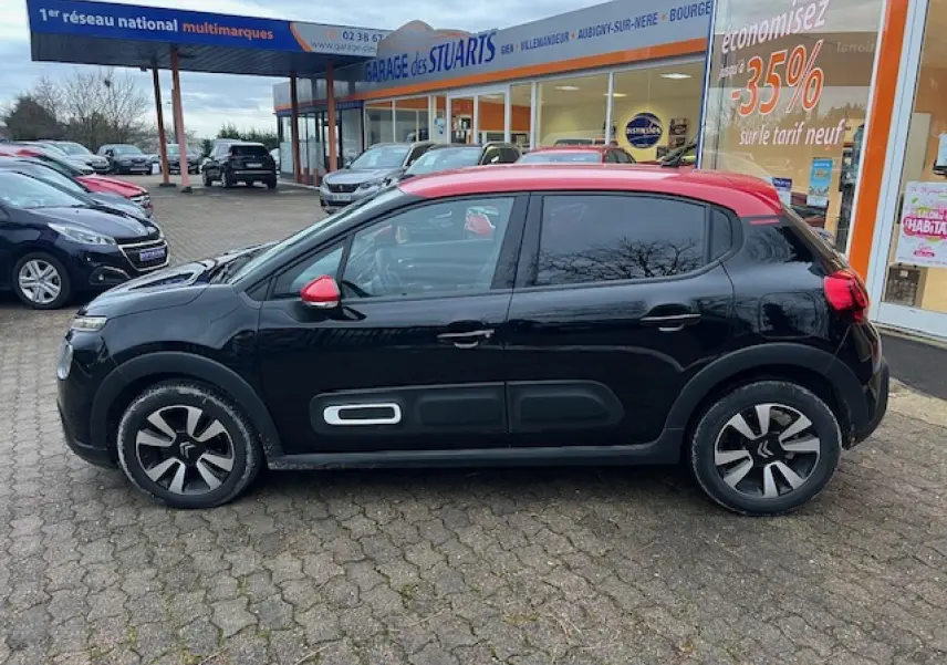 Profil côté gauche d'une Citroën C3 Shine 2023 noire avec toit rouge et jantes alliage diamantées, stationnée devant un garage.