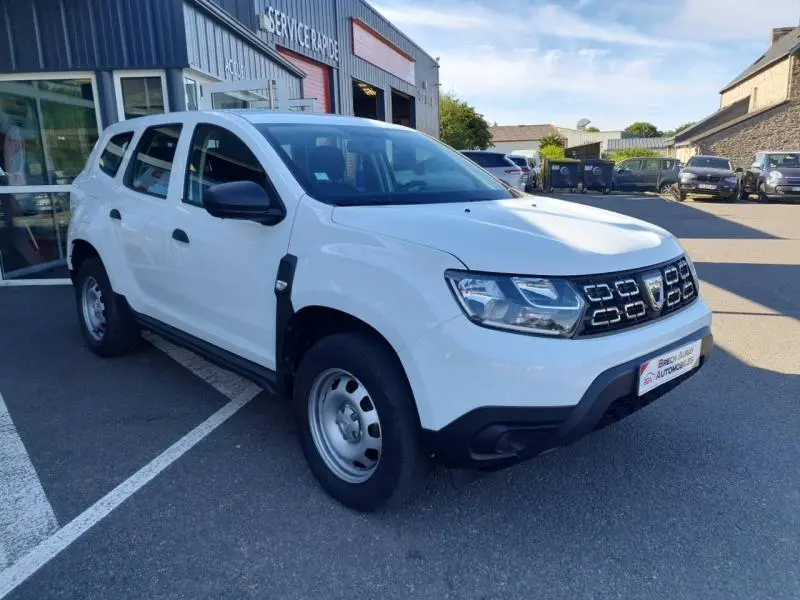 Dacia Duster blanc en 3/4 avant droit, avec calandre noire et jantes acier, stationné devant un garage.