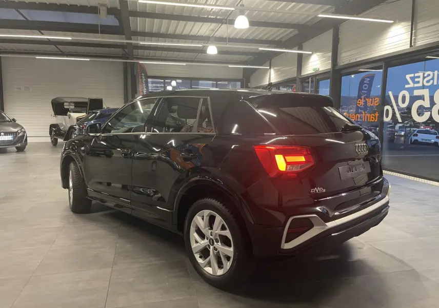 Audi Q2 noir métallisé vue 3/4 arrière droit en intérieur, feux arrière allumés et jantes en alliage visibles.