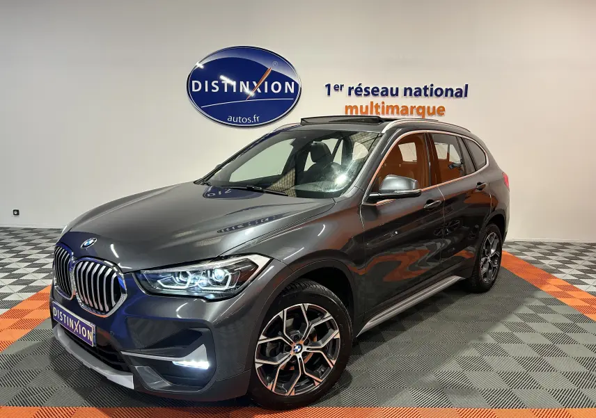 BMW X1 gris en vue 3/4 avant droit, avec toit ouvrant et jantes alliage distinctives, dans un showroom lumineux.