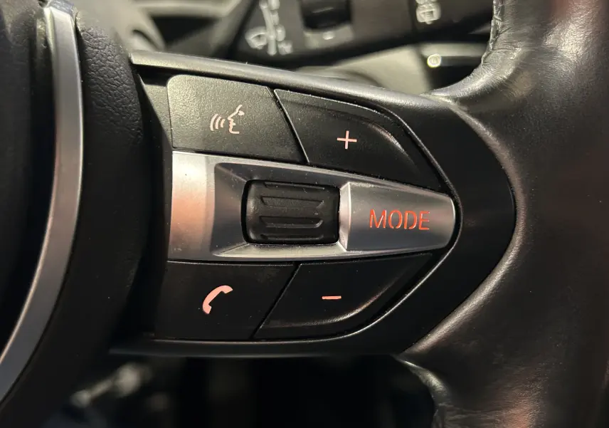 Gros plan sur les commandes au volant noir de la BMW X1 gris 2021, avec bouton mode éclairé en rouge.
