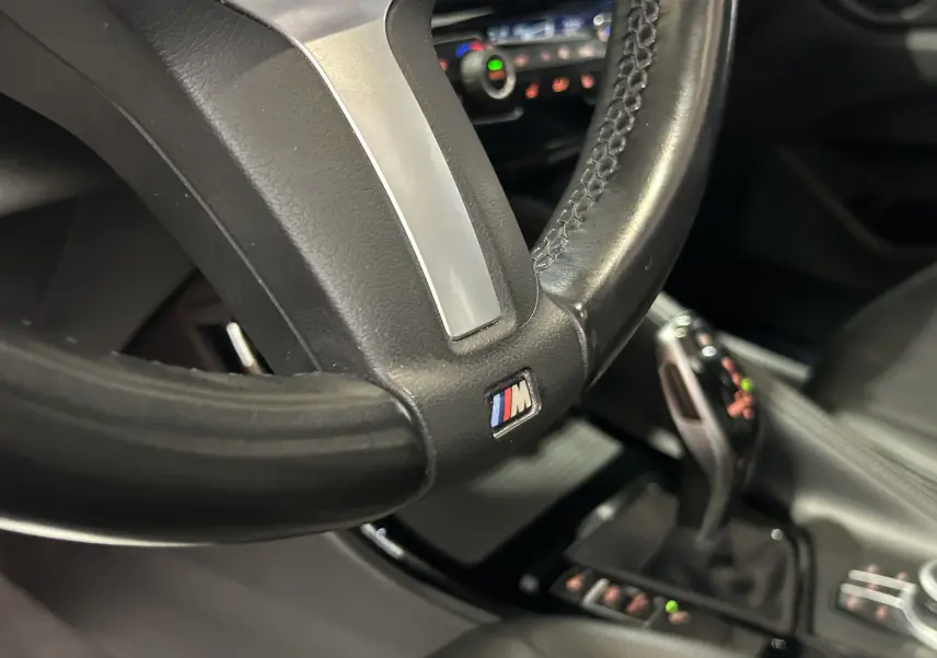 Gros plan sur le volant cuir avec logo M et levier de vitesse automatique dans l’habitacle noir du BMW X1 gris.