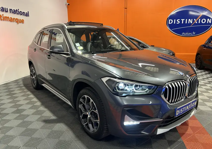 BMW X1 gris en 3/4 avant droit avec phares allumés et toit ouvrant visible en intérieur showroom.