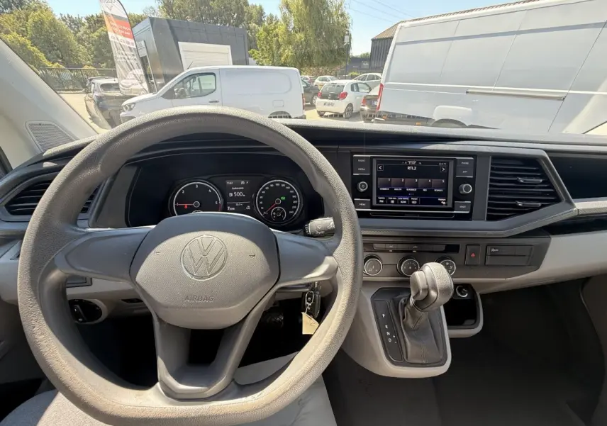 Vue intérieure centrée sur le volant et le tableau de bord du Volkswagen Transporter gris clair, avec écran tactile et levier de vitesses automatique.