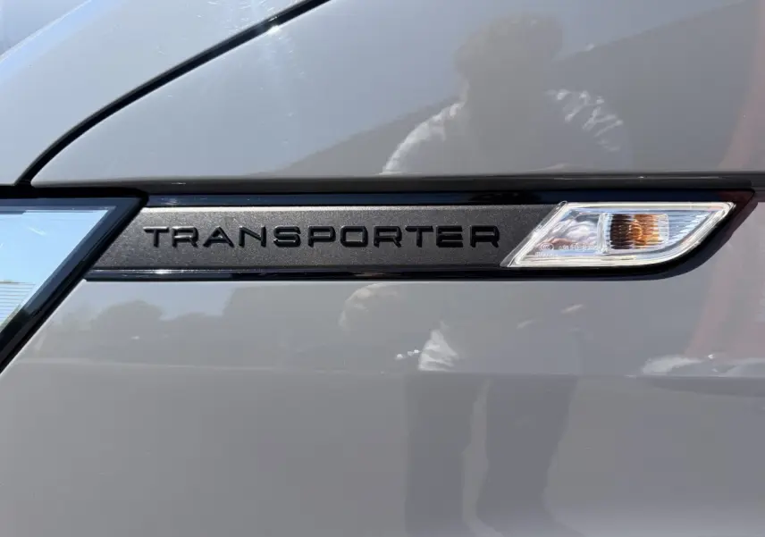 Détail du logo "Transporter" noir sur la porte gris clair d'un Volkswagen Transporter 2023, avec répétition lumineuse intégrée.
