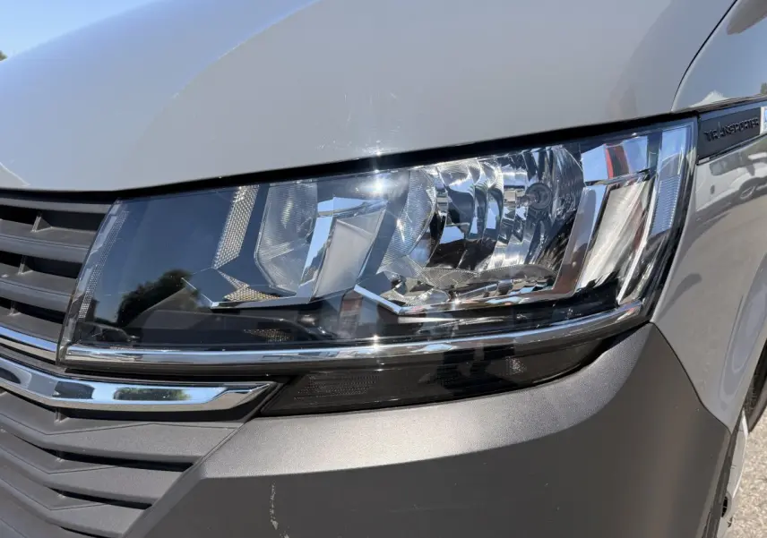 Gros plan sur le phare avant droit du Volkswagen Transporter gris clair, avec calandre et logo partiellement visibles.