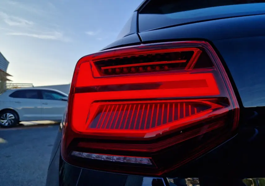 Gros plan sur le feu arrière droit rouge vif d’un Audi Q2 noir, avec un autre véhicule blanc en arrière-plan.