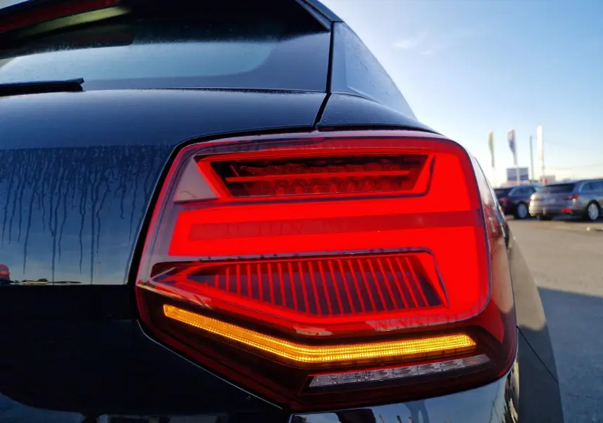 Gros plan sur le feu arrière droit rouge et orange d’un Audi Q2 noir, avec reflets et ciel bleu en arrière-plan.