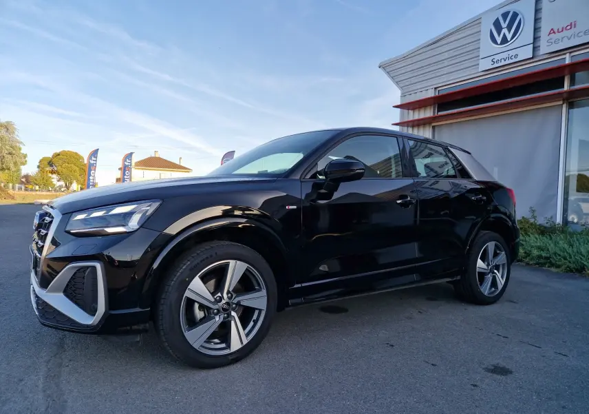 Audi Q2 noir en 3/4 avant droit, finition S line avec jantes alliage et calandre distinctive, stationné devant un service VW.