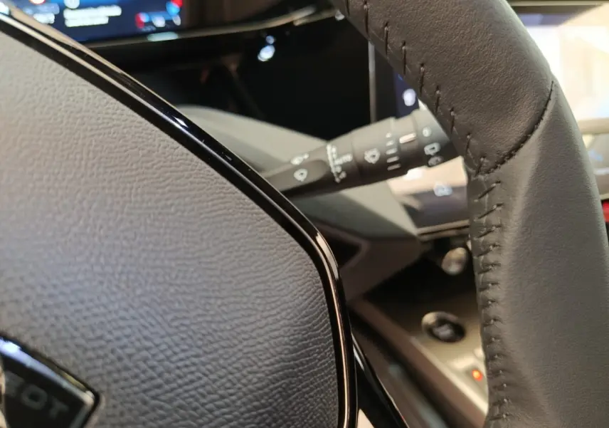 Gros plan sur le volant cuir noir multifonctions de la Peugeot 308 2026, avec tableau de bord et commandes visibles en arrière-plan.