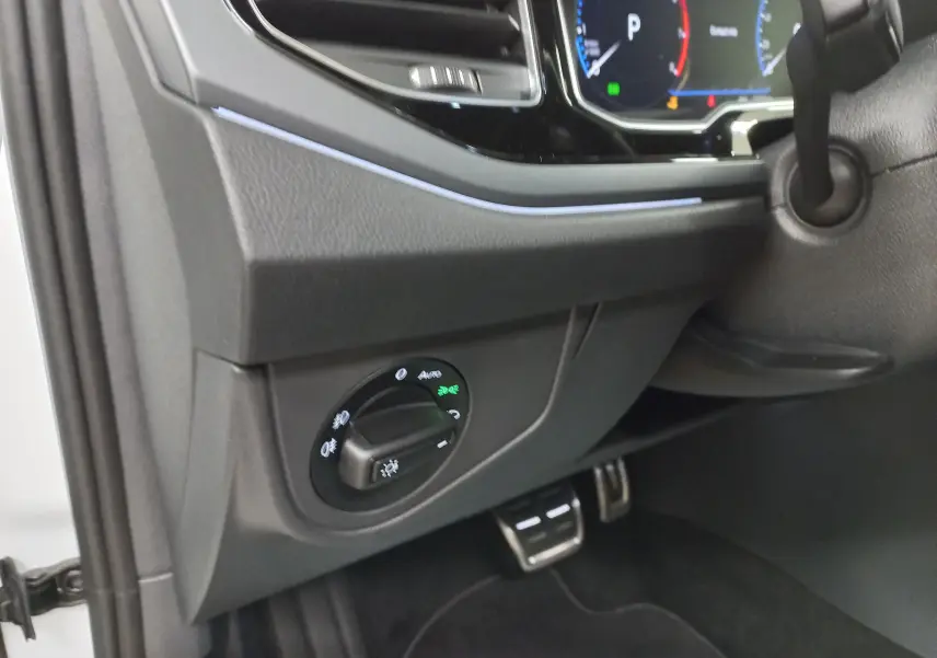 Détail intérieur côté gauche du tableau de bord du Volkswagen Taigo blanc 2025, avec commandes d'éclairage et pédales visibles.