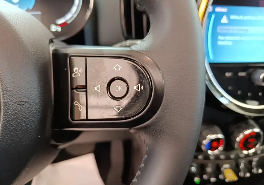 Gros plan sur le volant cuir noir multifonctions du MINI Countryman hybride, avec commandes vocales et écran tactile en arrière-plan.